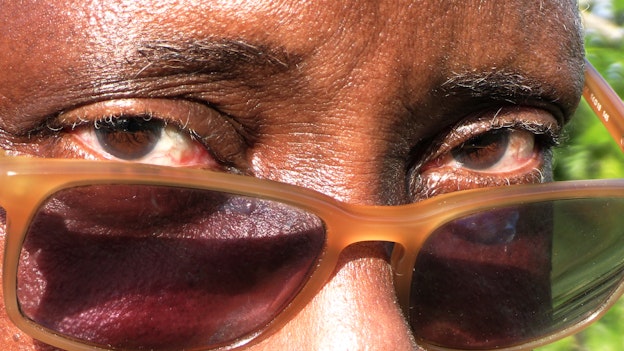 Video still of a close up of a persons face. Pictured are their eyes, which look into the camera, their eyebrows, and part of their nose. They are wearing sunglasses with a caramel, rectangular frame low on the bridge of their nose as the sun shines against their skin. There is greenery behind them.