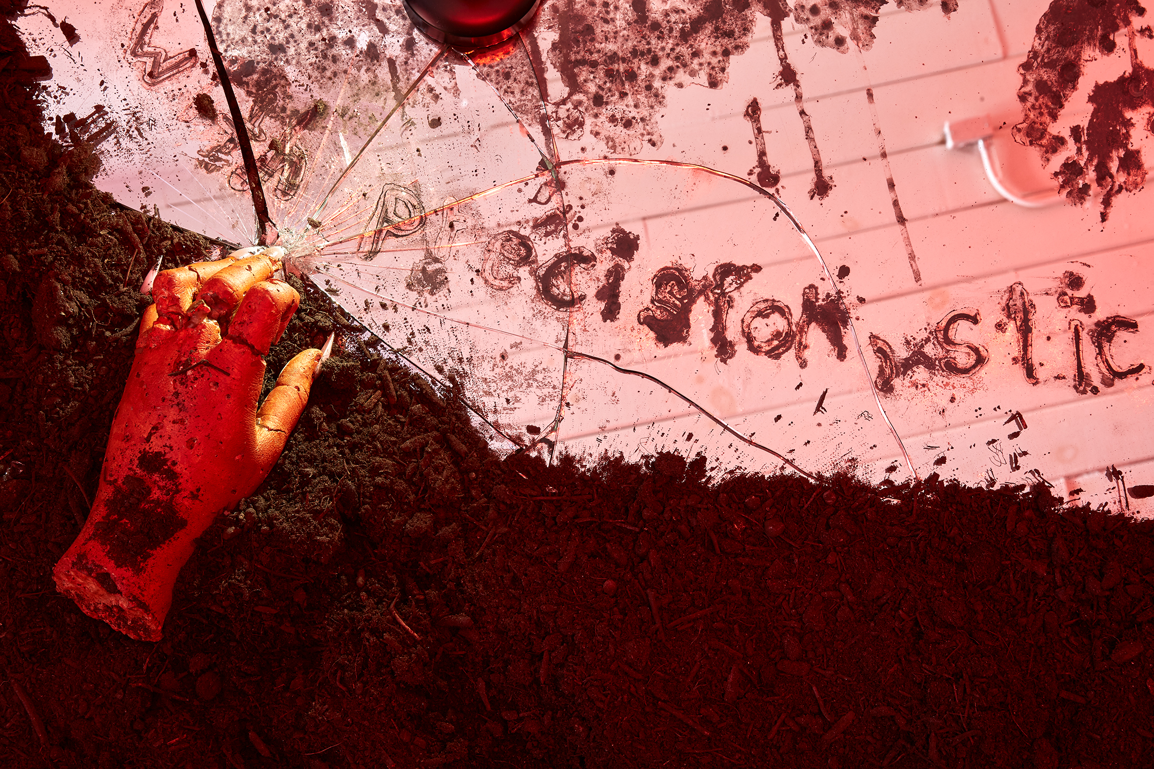 Red-lit installation view of a corner of the pool in ca(r)milla. The pool is made out of a mirror, which is bordered by soil. On top of the soil is a fake hand with long pink nails. The middle finger of the hand touches the edge of the pool, shattering a chunk of it. Fake blood is spread throughout the pool in both writing and blobs. The ceiling of the room reflects onto the pool.