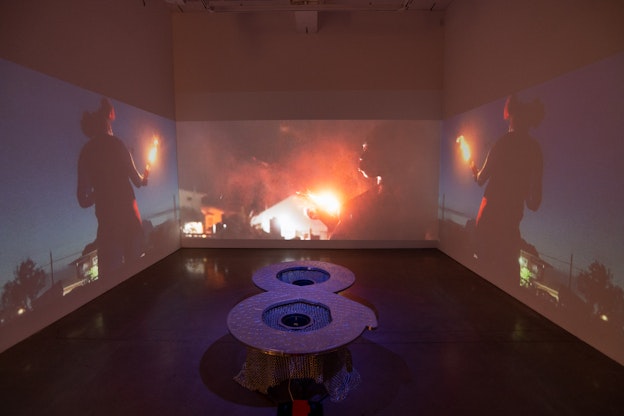 Installation view of “pappyshow in the dark time, my love”, an immersive gallery room with orange lighting. Three projections take up the width of the three white walls, with the left and right showing mirror images of a person’s silhouette, dancing in the night with a Molotov cocktail in their hand in front of a house. The center projection shows another angle of the dancer—outlining their face, the Molotov cocktail, and more houses in the night. In the center of the room, on the ground, is a sculptural object shaped like an “8”, made of aluminum and engraved with a pattern that resembles a snake’s skin. Inside of the two centers of the “8” are two subwoofers, each covered with chainmail in the shape of round table skirts.