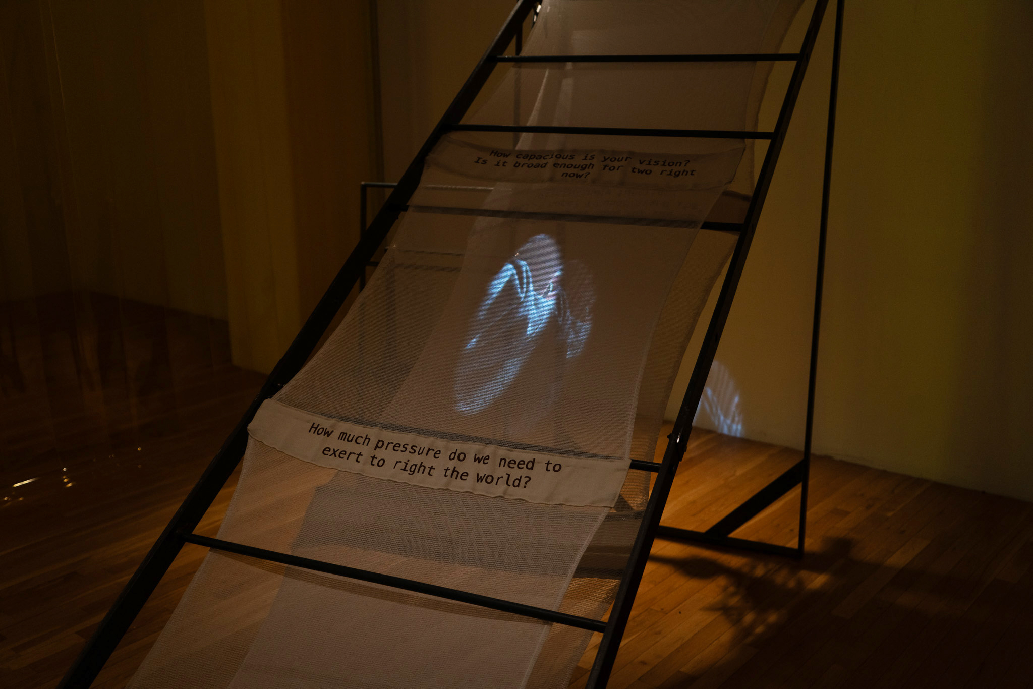 Installation view of “divinity surplus”, a black iron structure that resembles a ladder in a dimly lit gallery room with wooden floors. Scrim is casted through the body of the piece, going over and under the panels of the ‘ladder’. Two rectangles of brown cotton with black text printed onto it are threaded into the scrim. The scrim in the lower rectangle says “How much pressure do we need to exert to right the world?”, while the scrim placed higher reads “How capacious is your vision? Is it broad enough for two right now?”. Two ovals with light blue and grey textures are projected onto the center of the scrim, which bleeds through it onto the white wall. Sheer black curtains hang to the left of the installation.