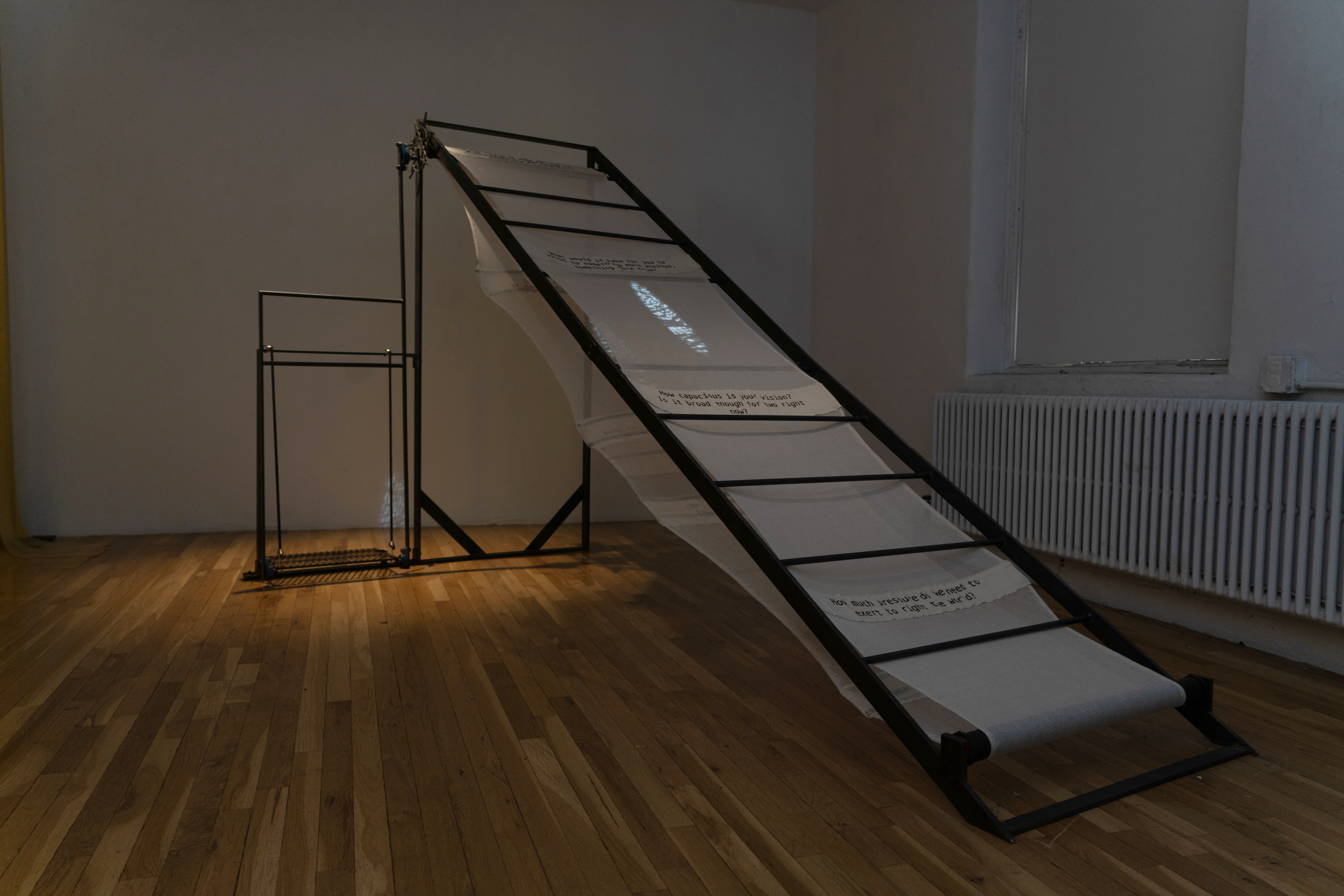 Installation view of “divinity surplus”, a black iron structure that resembles a ladder in a dimly lit gallery room with wooden floors. Scrim is casted through the body of the piece, going over and under the panels of the ‘ladder’. Four rectangles of brown cotton with printed black text are threaded into the scrim in nearly even intervals. The text is illegible due to the distance from which the photo was taken. An elongated blue oval with grey textures is projected onto the center of the scrim, which bleeds through it onto the white wall.