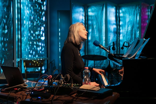 Olivia Block mid-performance. She is sitting down in front of a piano that she is playing with one hand while singing into a microphone that hangs in front of her sheet music. There is a table of electronic equipment with a red beverage in a plastic bottle laid out in front of her. Wires are plugged into and coming out of her gear. Behind Block are room dividers, panels, and a bit of a wall that is lit up with solid blue, sea green, and purple projections, as well as white dots and white amorphous shapes. She is wearing a black turtleneck with a gold necklace over it.