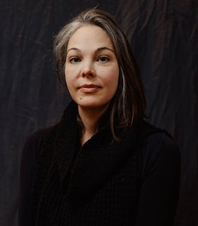 Headshot of Olivia Block looking directly at the camera. She is wearing a black shirt and a black vest. Behind her is dark fig color backdrop with wrinkles.
