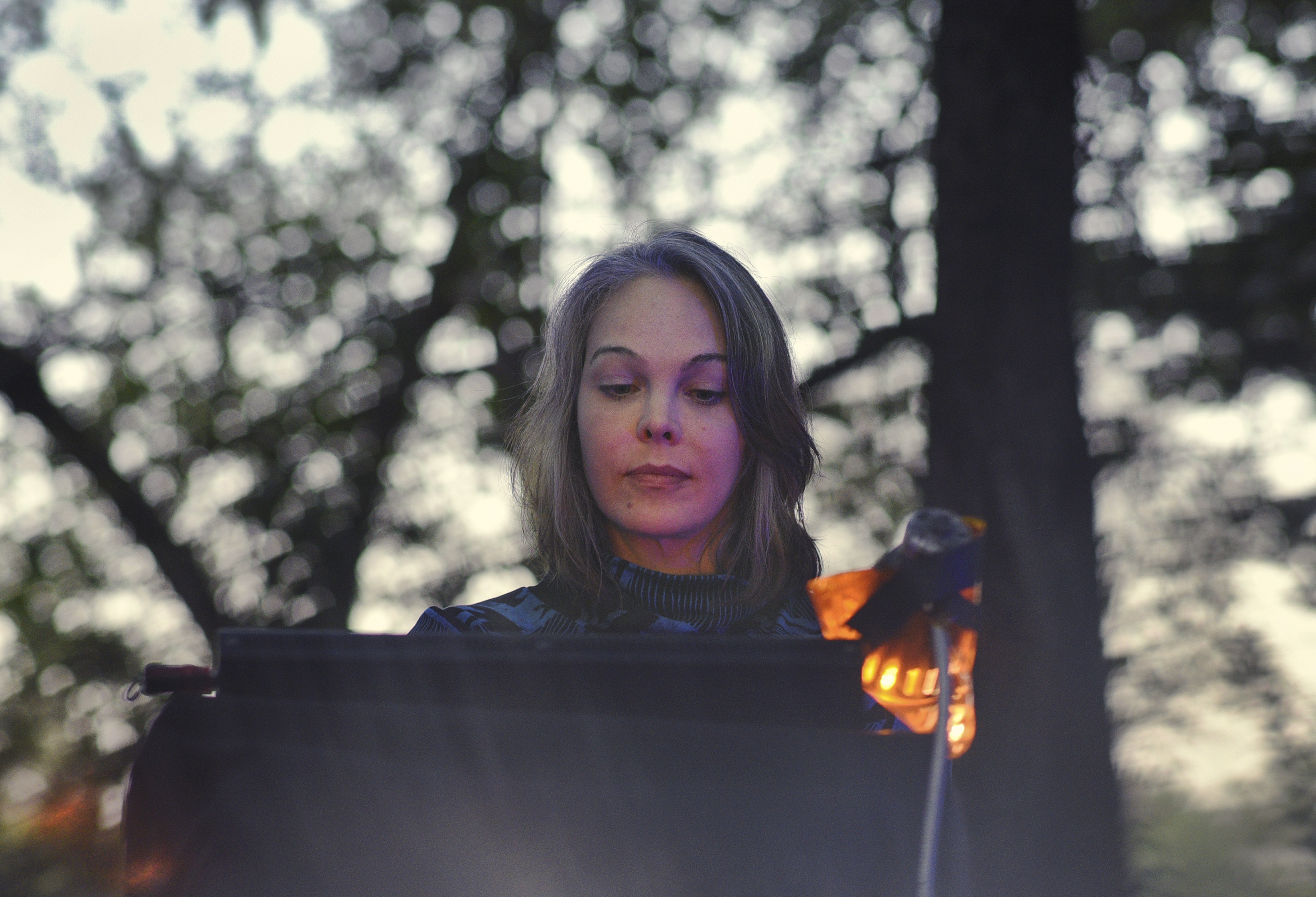Olivia Block looking down at her electronic equipment during an outdoor performance. To the right of Block is a flexible gooseneck lamp with an orange lightbulb. She is wearing a blue and black patterned turtleneck. Behind her are blurry trees.