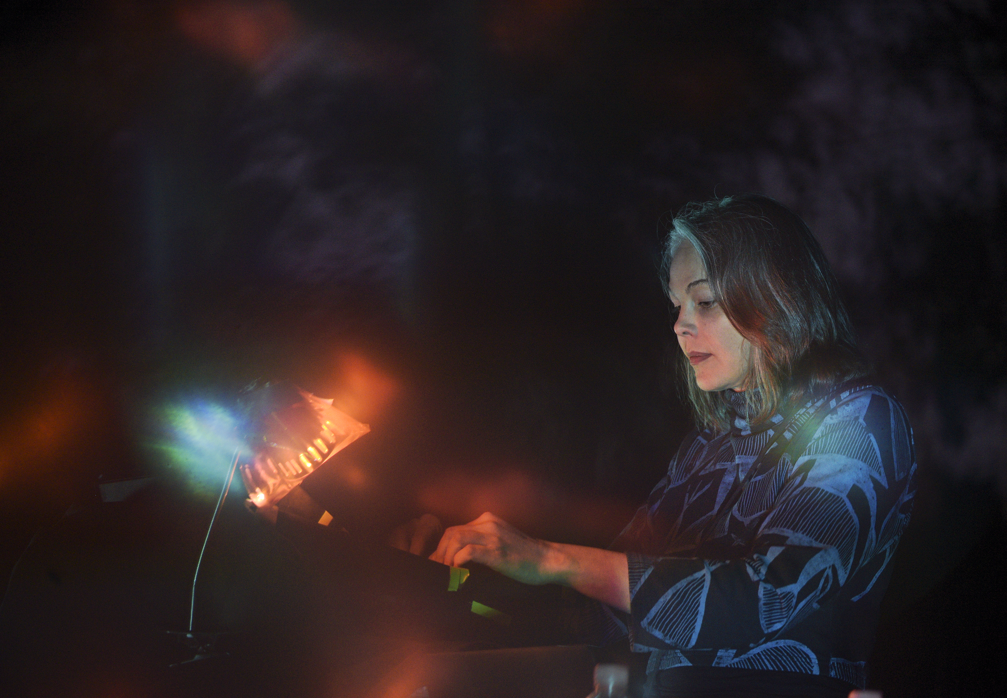 A front-side view of Olivia Block performing outdoors. She is looking down as her hands move over her electronic music equipment, which has a few green tape strips on its side and is illuminated by a flexible gooseneck lamp with an orange lightbulb. There are some blue and green lights hovering above her gear that seem to be originating in a synthesizer and then filtering through through the flash of the photograph. The orange lights emulate blobs and appear throughout. Block is wearing a black and blue patterned turtleneck, which foreshadows the blurry trees in the background.