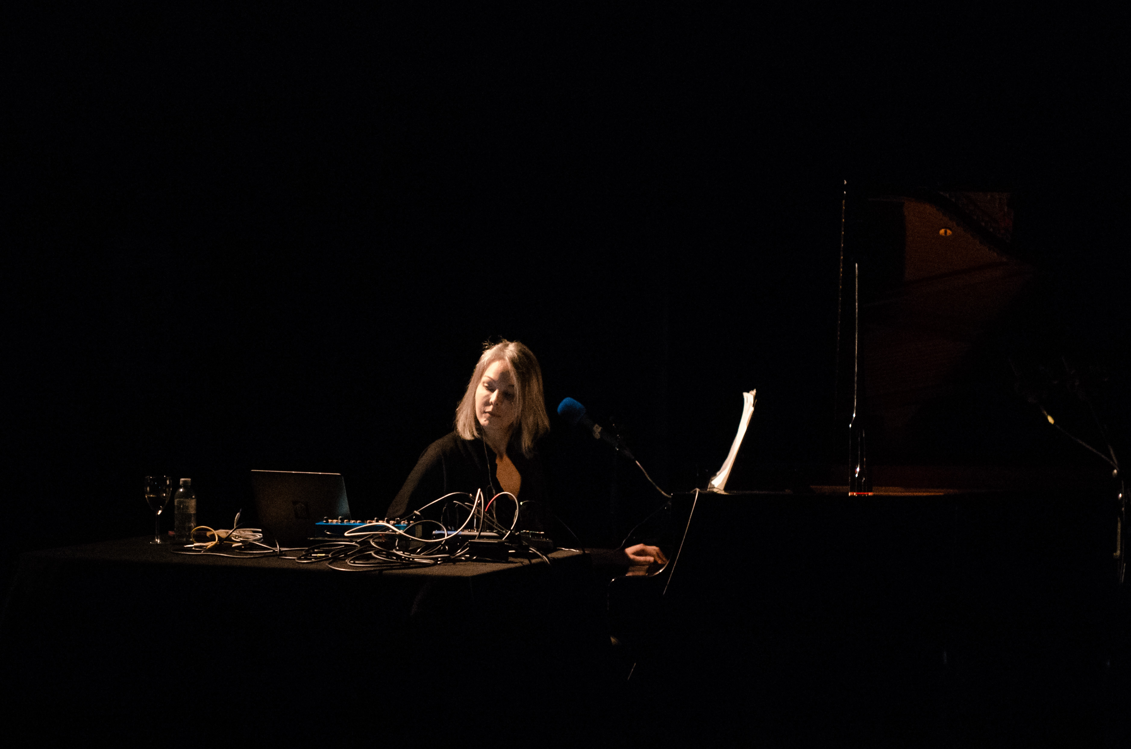 Olivia Block is sitting in a (dark) room. There is a stage light directed at her as she looks down onto her table of electronic equipment, which has long cables going in and out of the gear, along with a plastic water bottle and a wine glass. To the right side is a piano with sheet music and a clipped vocal microphone.