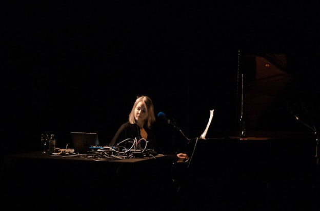 Olivia Block is sitting in a (dark) room. There is a stage light directed at her as she looks down onto her table of electronic equipment, which has long cables going in and out of the gear, along with a plastic water bottle and a wine glass. To the right side is a piano with sheet music and a clipped vocal microphone.