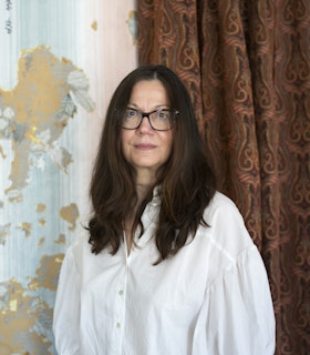 Headshot of Brenda Coultas. Coultas is looking at the camera, wearing a white long sleeve button-up collared shirt. Her body is positioned between the two different materials on the wall behind her. To Coultas' right is a patterned curtain in autumn colors with a bit of baby blue. To her left is wallpaper with beige, white, seafoam blue and baby blue lines running up and down, separated by color, and interrupted by dark yellow and blue shapes.