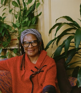 Portrait of Nanette Carter in a pastel yellow room. Smiling at the camera, Carter is wearing a fuzzy orange sweater with black sleeves and a black and white striped head wrap. She is sitting on a section of a couch that is orange with a blue, pink, and yellow pattern throughout it. Behind Carter is another section of the couch, but this time it is in brown, with a small orange pillow with pink, blue, yellow, and red patterns on it. Three vibrant green plants are around Carter. 