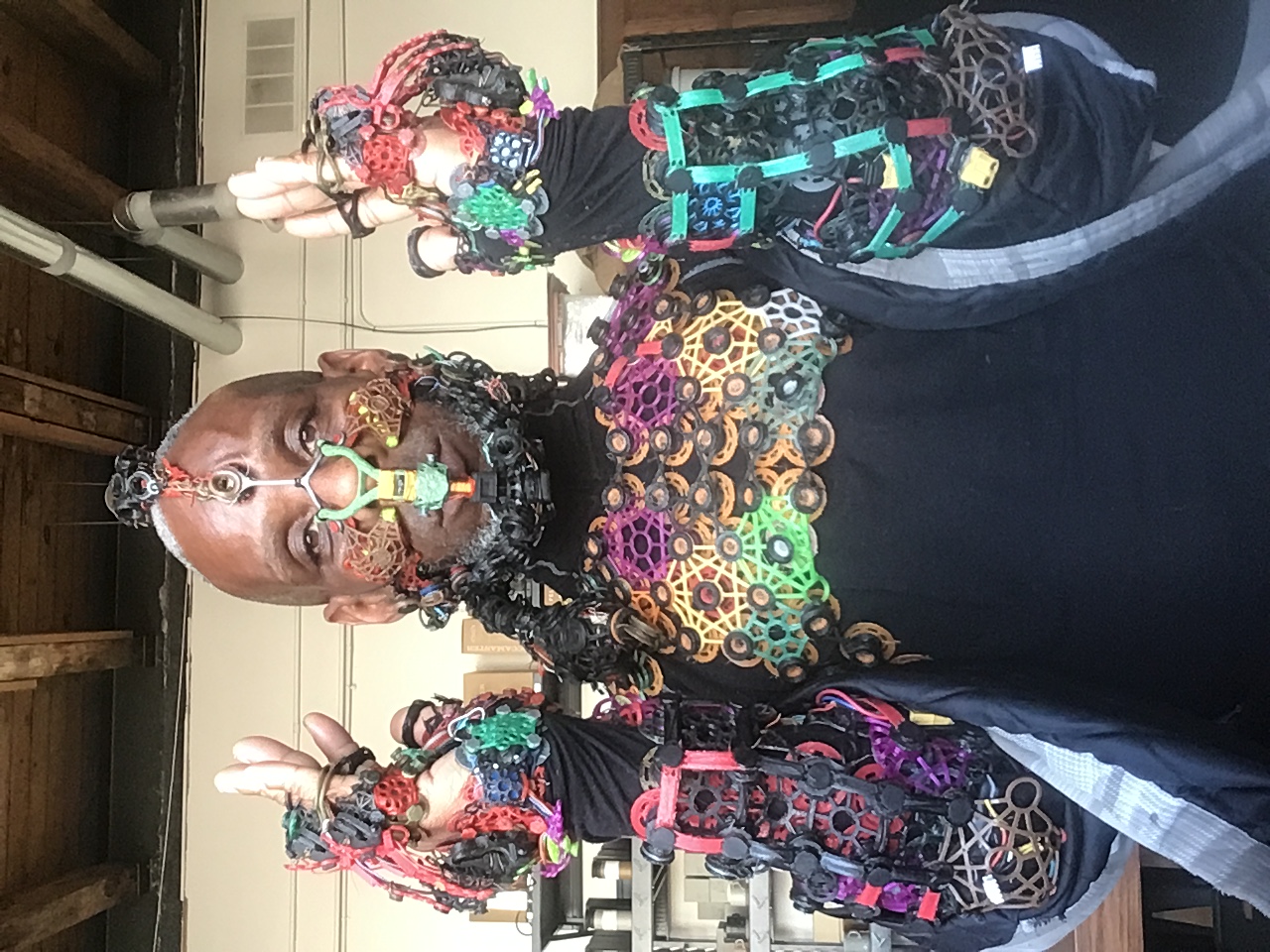 Headshot of Onyx Ashanti in a studio, looking at the camera with their hands by their ears as they display their multi-colored exodermal mesh interface over the top half of their body. The interface begins at the top center of their head, going down their forehead to their nose, at which point it becomes symmetrical. It also rests on their cheeks, lips, beard, chest, forearms, and hands.