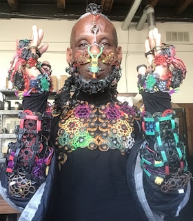 Headshot of Onyx Ashanti in a studio, looking at the camera with their hands by their ears as they display their multi-colored exodermal mesh interface over the top half of their body. The interface begins at the top center of their head, going down their forehead to their nose, at which point it becomes symmetrical. It also rests on their cheeks, lips, beard, chest, forearms, and hands.