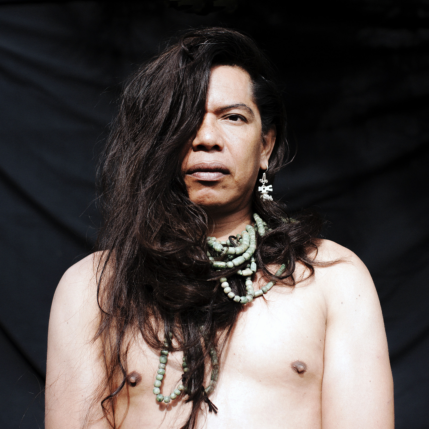 Headshot of Lukas Avendaño. Avendaño looks at the camera, her hair is covering her left eye  and folded into various parts of the beaded turquoise necklace wrapped around her neck  in multiple layers. She is wearing a dangling silver earring in her right ear and is shirtless. There is a black backdrop behind Avendaño.