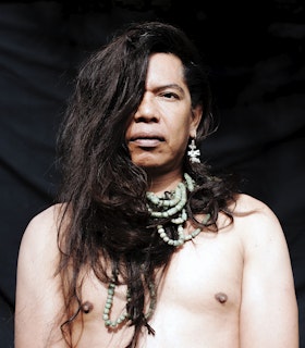 Headshot of Lukas Avendaño. Avendaño looks at the camera, her hair is covering her left eye  and folded into various parts of the beaded turquoise necklace wrapped around her neck  in multiple layers. She is wearing a dangling silver earring in her right ear and is shirtless. There is a black backdrop behind Avendaño.