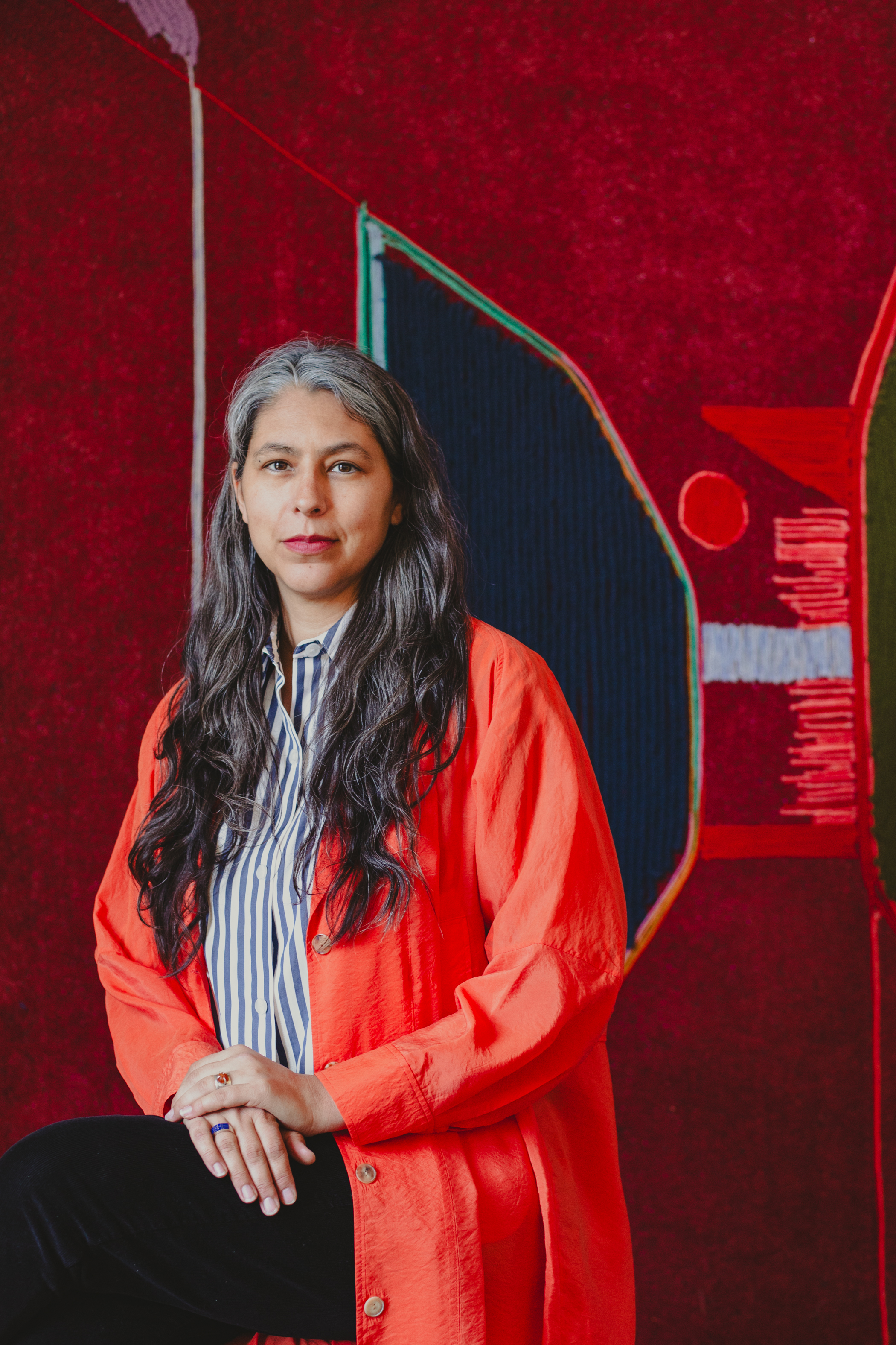 Portrait of Teresa Baker. Baker smiles at the camera, sitting with her hands placed on top of each other resting over her crossed leg. She’s wearing black pants, a blue and white striped button up shirt with a bright orange jacket over it, and silver rings on each ring finger, one with lapis lazuli and the other a stone that resembles sunstone or amber.  Behind Baker her work “Twenty Minutes to Sunset” appears in close detail.