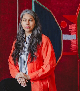 Portrait of Teresa Baker. Baker smiles at the camera, sitting with her hands placed on top of each other resting over her crossed leg. She’s wearing black pants, a blue and white striped button up shirt with a bright orange jacket over it, and silver rings on each ring finger, one with lapis lazuli and the other a stone that resembles sunstone or amber.  Behind Baker her work “Twenty Minutes to Sunset” appears in close detail.