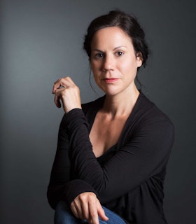 Portrait of Jennifer Elise Foerster wearing a black long  sleeve top and blue jeans. She is looking at the camera while sitting down with her right leg up,  which she is  resting her left hand on as well as her right elbow.  Foerster's right hand is in the air just over her left  shoulder. There is a grey backdrop behind her.