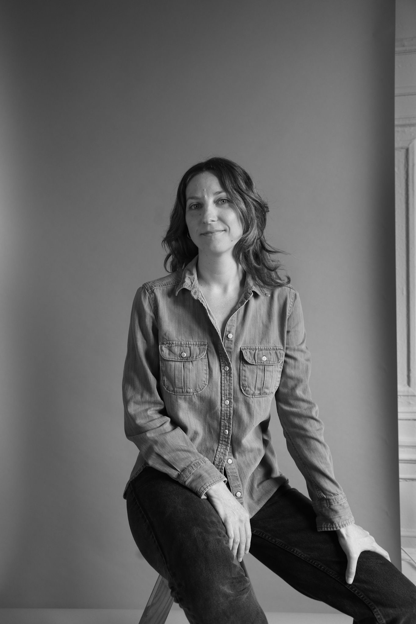 Black and white portrait of Maggie Cloud. She is sitting on a wooden ladder,  smiling at the camera in jeans and a sleeveless fringe shirt. Maggie’s feet are  bare, pointed down on the last step of the ladder and the floor. Behind her is a  white backdrop.