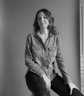 Black and white portrait of Maggie Cloud. She is sitting on a wooden ladder,  smiling at the camera in jeans and a sleeveless fringe shirt. Maggie’s feet are  bare, pointed down on the last step of the ladder and the floor. Behind her is a  white backdrop.