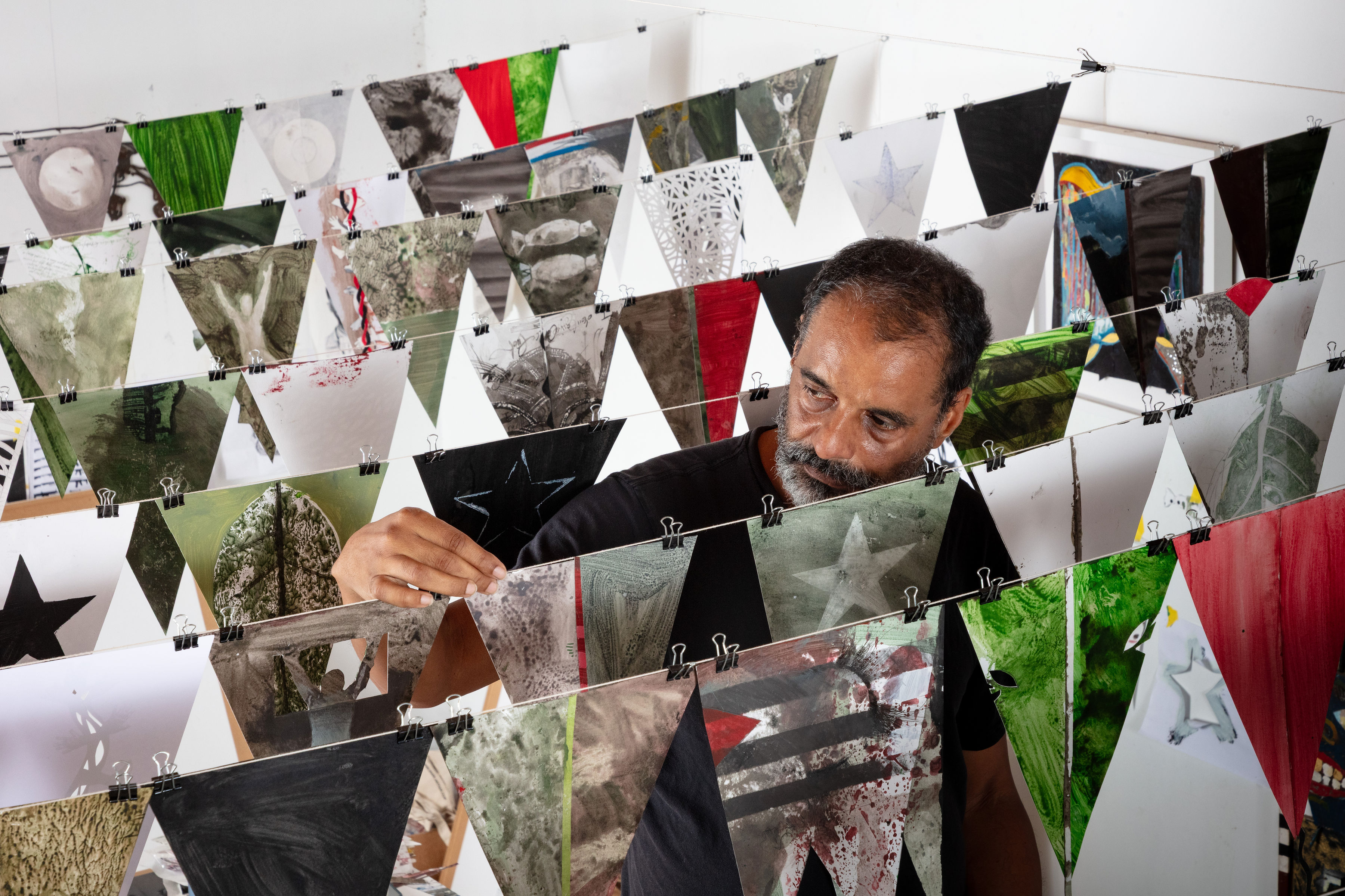 Portrait of Christopher Cozier adjusting a row of pennant string flags in a gallery space. There are 7 rows in total, with each flag painted with different images in what are varying earth tones, in addition to red and bright green. Images on the flags include varying stars, candy wrappers, leafs, the moon, and what resembles the Cuban flag, minus the star. Each flag has two binder clips on it, attaching it to a white string. The flags are installed against white gallery walls, above a white floor. On the wall behind Christopher is a framed artwork.