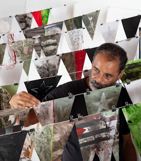 Portrait of Christopher Cozier adjusting a row of pennant string flags in a gallery space. There are 7 rows in total, with each flag painted with different images in what are varying earth tones, in addition to red and bright green. Images on the flags include varying stars, candy wrappers, leafs, the moon, and what resembles the Cuban flag, minus the star. Each flag has two binder clips on it, attaching it to a white string. The flags are installed against white gallery walls, above a white floor. On the wall behind Christopher is a framed artwork.