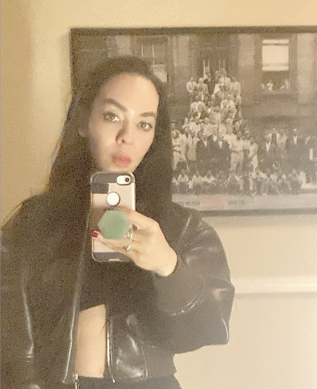 Grainy mirror selfie of Harmony Holiday. Holiday is looking directly at the mirror, covering her neck with her phone in the reflection. She’s holding her phone with a green hexagonal phone grip attached to her light pink phone case. She is wearing an unzipped leather jacket, which exposes part of her stomach. There is a yellow wall behind Holiday as well as a framed 
