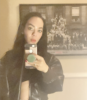 Grainy mirror selfie of Harmony Holiday. Holiday is looking directly at the mirror, covering her neck with her phone in the reflection. She’s holding her phone with a green hexagonal phone grip attached to her light pink phone case. She is wearing an unzipped leather jacket, which exposes part of her stomach. There is a yellow wall behind Holiday as well as a framed 