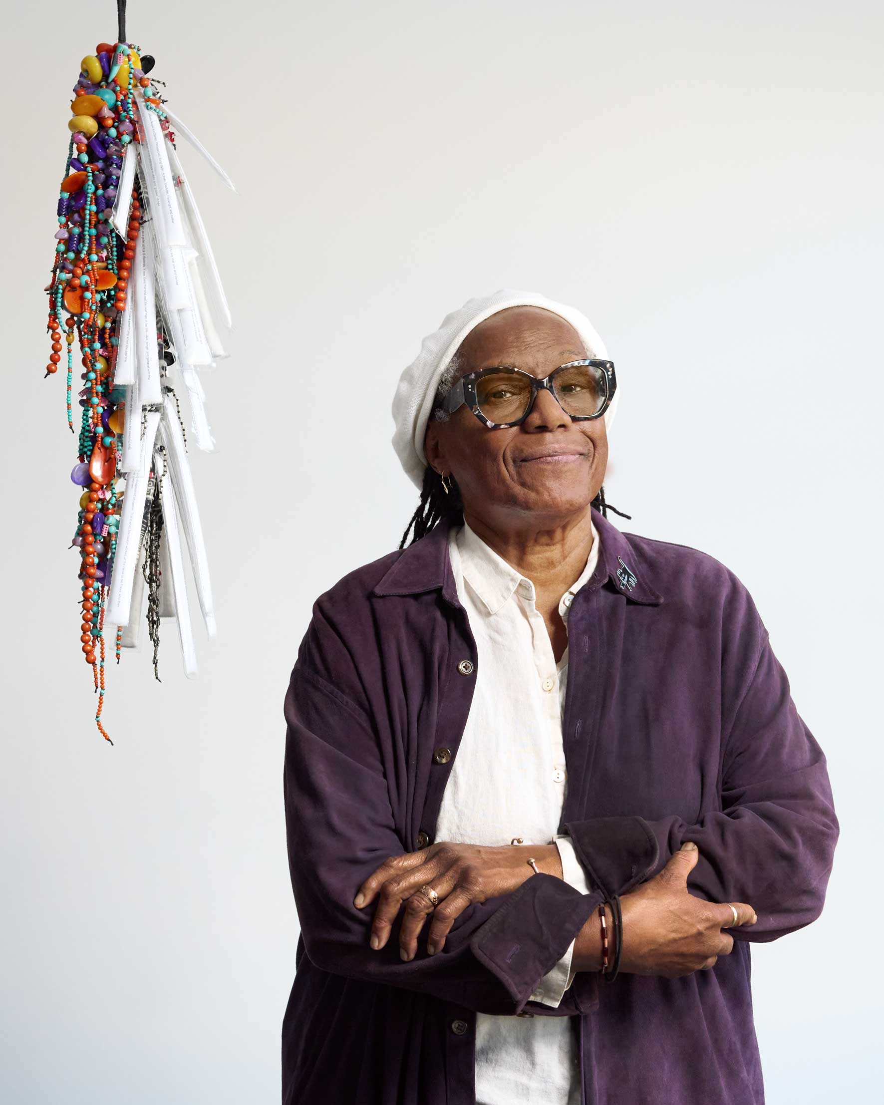 Portrait of Janet Olivia Henry looking at the camera and holding her arms across  her chest, wearing a suede purple button down jacket over a white collard shirt,  a white hat, as well as jewelry on her wrists, fingers and ear. Henry’s jacket is  decorated with a pin of an animated black cat on its collar. Behind and to the  left of Henry is a dangling sculpture composed of beads.