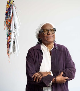 Portrait of Janet Olivia Henry looking at the camera and holding her arms across  her chest, wearing a suede purple button down jacket over a white collard shirt,  a white hat, as well as jewelry on her wrists, fingers and ear. Henry’s jacket is  decorated with a pin of an animated black cat on its collar. Behind and to the  left of Henry is a dangling sculpture composed of beads.