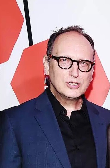 Headshot of Paul Lazar. Lazar looks at the camera, smiling with his teeth. He’s wearing a blue suit with a black button up shirt underneath. Lazar appears to be on a red carpet or at corporate event entrance, standing  in front of a white backdrop that includes two red shapes and a black line. The image is cropped just over his head and below his chest, presumably cutting others who were originally in the photo with him. 