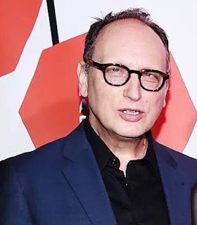 Headshot of Paul Lazar. Lazar looks at the camera, smiling with his teeth. He’s wearing a blue suit with a black button up shirt underneath. Lazar appears to be on a red carpet or at corporate event entrance, standing  in front of a white backdrop that includes two red shapes and a black line. The image is cropped just over his head and below his chest, presumably cutting others who were originally in the photo with him. 