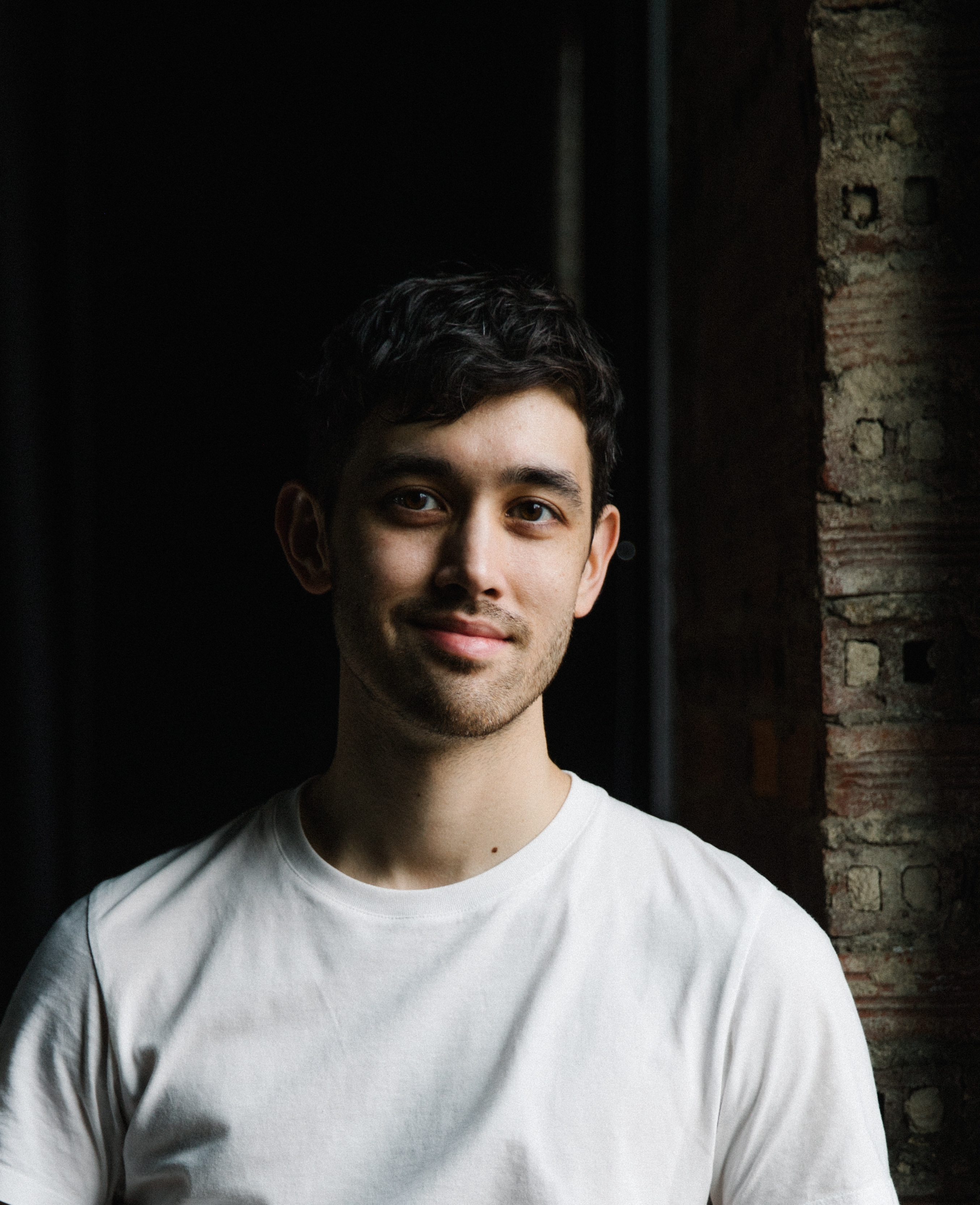 Headshot of Benjamin Akio Kimitch. Looking at the camera, he smiles to his right side.  Kimitch is wearing a white shirt and is in front of a black backdrop.