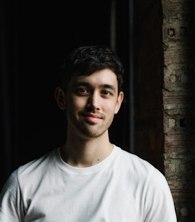 Headshot of Benjamin Akio Kimitch. Looking at the camera, he smiles to his right side.  Kimitch is wearing a white shirt and is in front of a black backdrop.