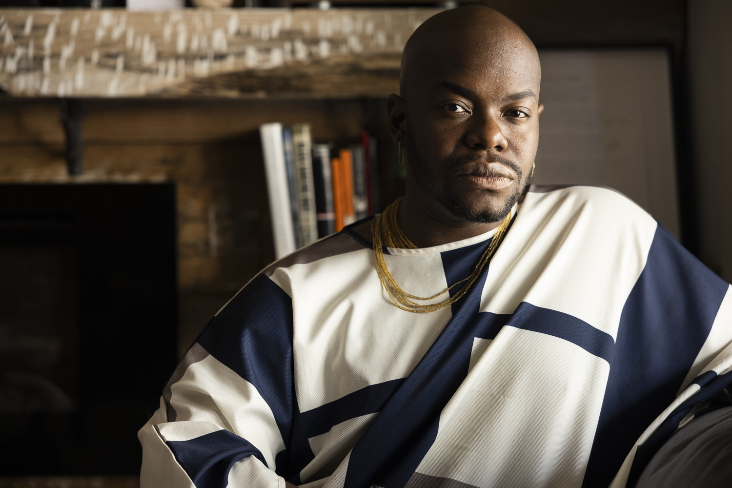 jaamil olawale kosoko sits outside on a large rock, holding their hands in a triangle, looking towards the camera. The sun is shining  over jaamil. They are wearing a black short sleeve shirt, black  pants, and a square turquoise and gold ring. Above the rock are various green plants, also basking in the sun. 