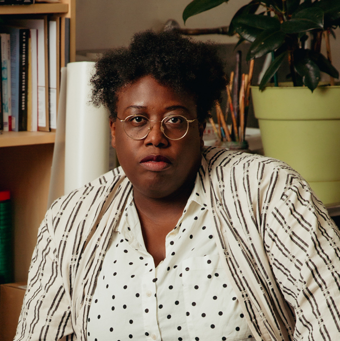 Portrait of Nyeema Morgan in her studio. Morgan is looking at the camera,  wearing a white dress shirt with black polkadots with an off-white patterned  cardigan on top. In particular, the pattern consists of black double lines that  go up and down, occasionally layered with beige double lines going in the  opposite direction. This creates an optical illusion---allowing the cardigan to  seem like it has black polkadots throughout too. Behind Morgan, from  left to right, is a bookshelf filled with books, a paper roll, lots of used paint  brushes in a jar, and a plant in a plastic green pot. 