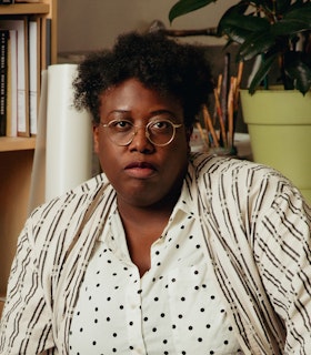 Portrait of Nyeema Morgan in her studio. Morgan is looking at the camera,  wearing a white dress shirt with black polkadots with an off-white patterned  cardigan on top. In particular, the pattern consists of black double lines that  go up and down, occasionally layered with beige double lines going in the  opposite direction. This creates an optical illusion---allowing the cardigan to  seem like it has black polkadots throughout too. Behind Morgan, from  left to right, is a bookshelf filled with books, a paper roll, lots of used paint  brushes in a jar, and a plant in a plastic green pot. 