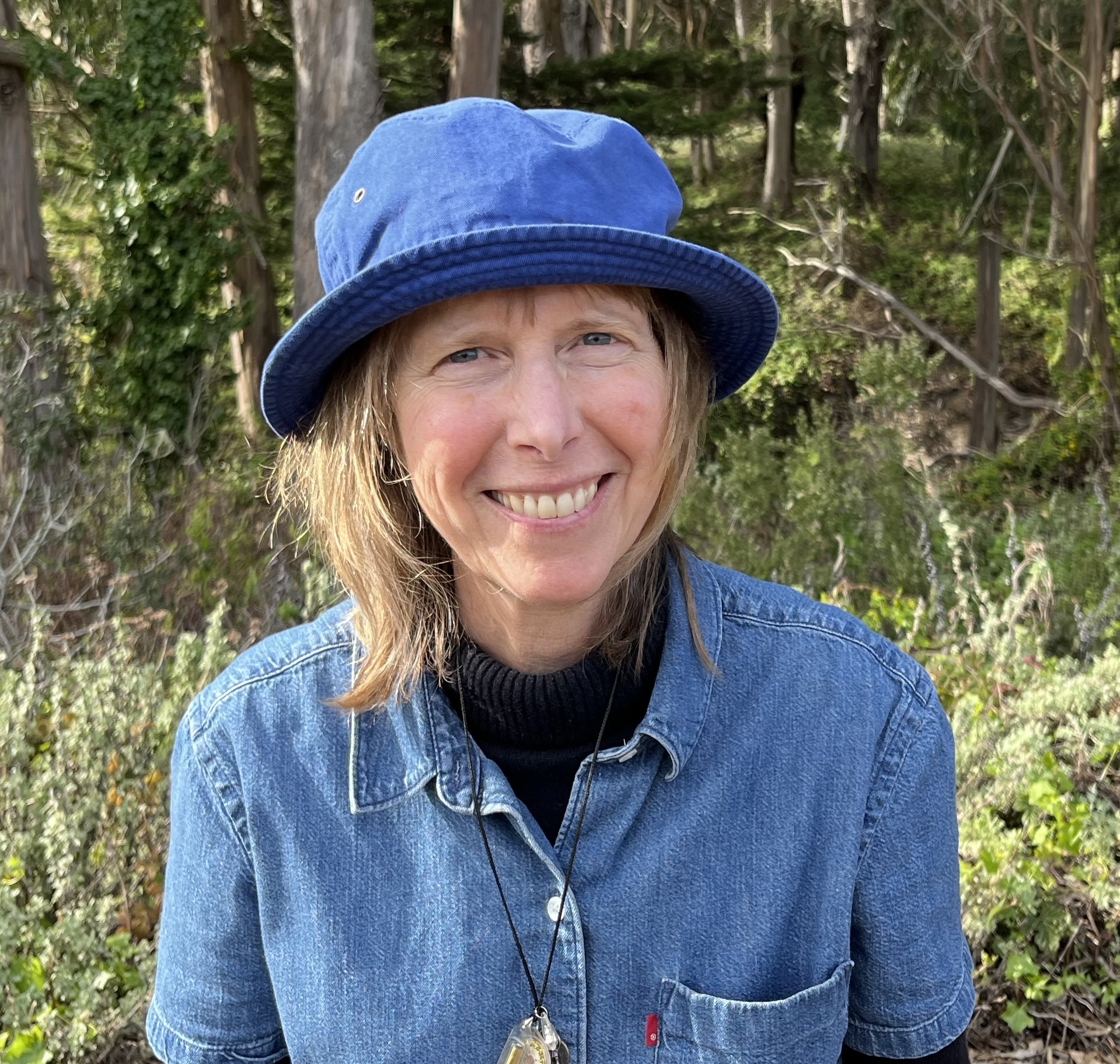 Headshot of Denise Newman in the woods. Newman is smiling at the camera, wearing a bright blue bucket hat, and a denim collared t-shirt over a black turtleneck. Her necklace hangs over her clothing. The sun is shining on the greenery and trees behind her.