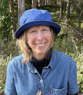 Headshot of Denise Newman in the woods. Newman is smiling at the camera, wearing a bright blue bucket hat, and a denim collared t-shirt over a black turtleneck. Her necklace hangs over her clothing. The sun is shining on the greenery and trees behind her.