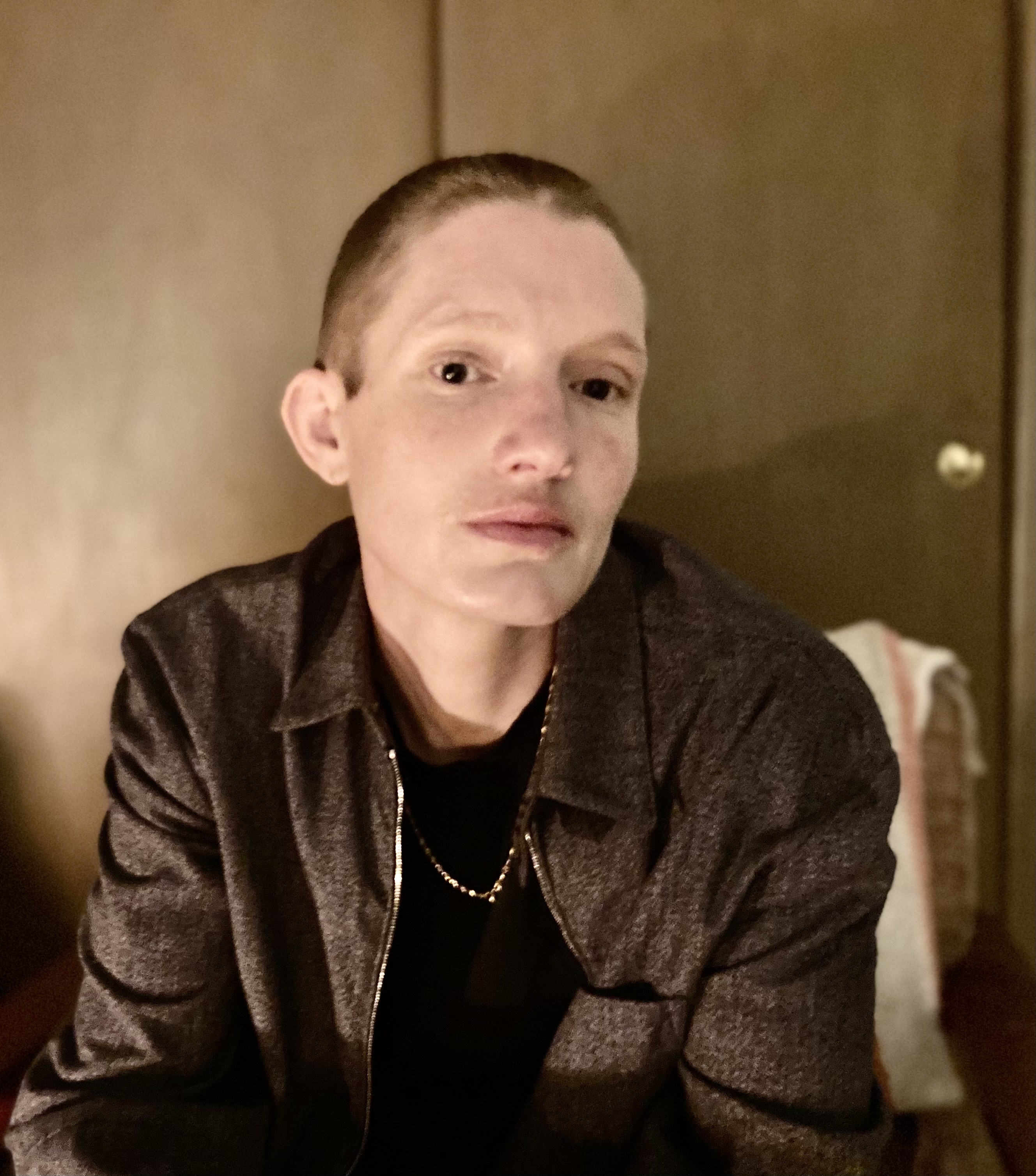 Portrait of Timmy Straw. Straw is sitting down and looking towards  the camera, wearing an unzipped grey collared jacket with a black  shirt and a thin gold chain. They are in front of a greenish brown  door. At the top and bottom of the portrait there is a pinkish grey  border containing white text. The top reads 