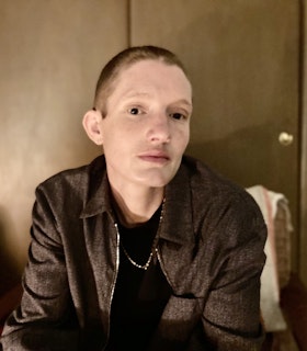 Portrait of Timmy Straw. Straw is sitting down and looking towards  the camera, wearing an unzipped grey collared jacket with a black  shirt and a thin gold chain. They are in front of a greenish brown  door. At the top and bottom of the portrait there is a pinkish grey  border containing white text. The top reads 