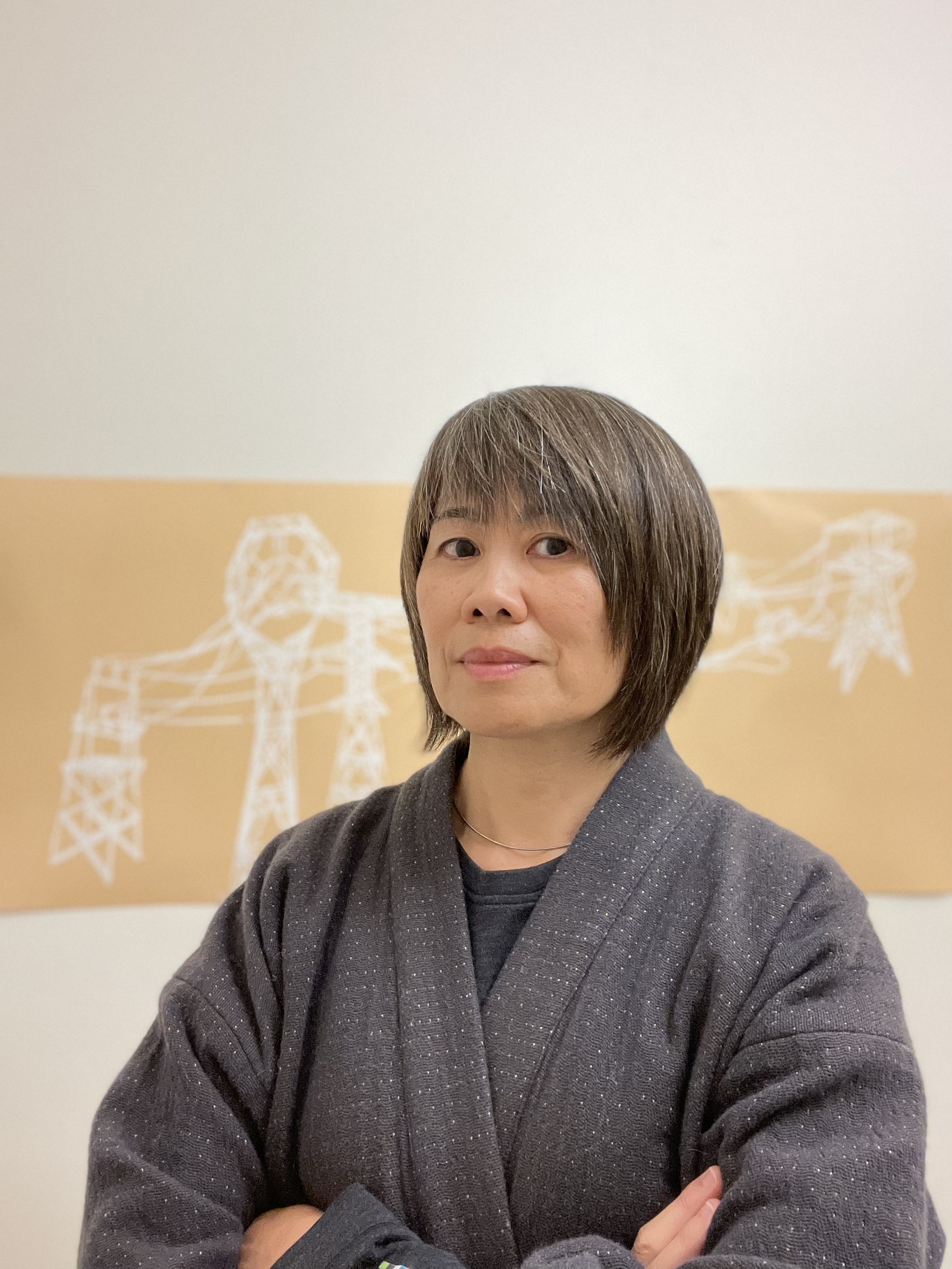 Portrait of Shirley Tse in front of an installation on a white gallery wall. Tse is looking at the  camera and crossing her arms, wearing a light gray cardigan with little white dots throughout   over a dark gray shirt. She is also wearing a gold necklace. The installation behind her  has a beige background with white drawings that resemble power lines.