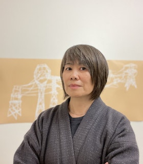 Portrait of Shirley Tse in front of an installation on a white gallery wall. Tse is looking at the  camera and crossing her arms, wearing a light gray cardigan with little white dots throughout   over a dark gray shirt. She is also wearing a gold necklace. The installation behind her  has a beige background with white drawings that resemble power lines.