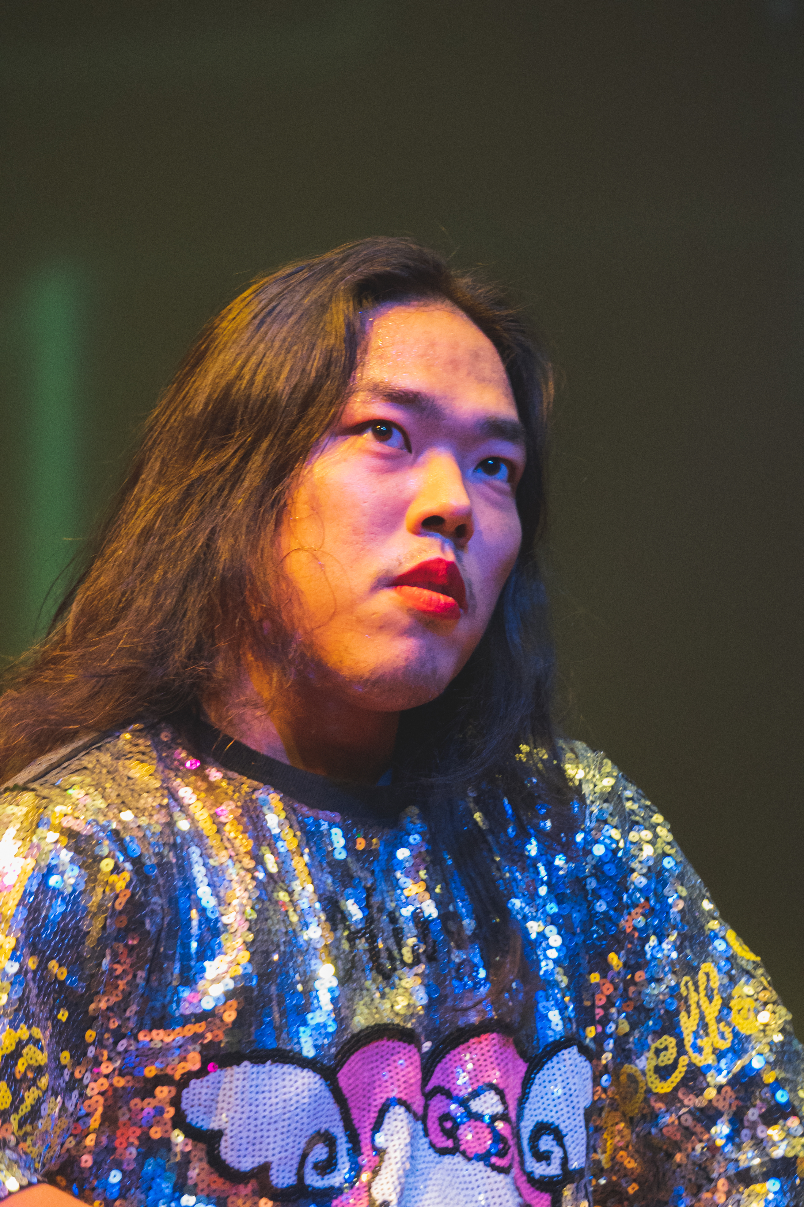 Anh Vo gestures forward with one hand, captured  mid-performance with their mouth open. They are wearing a white fur vest over a long sleeve black  dress with printed gold chains running up and down.  The room is dimly lit, creating shadows and a purple hue  in the background.