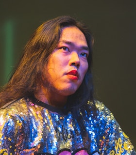 Anh Vo gestures forward with one hand, captured  mid-performance with their mouth open. They are wearing a white fur vest over a long sleeve black  dress with printed gold chains running up and down.  The room is dimly lit, creating shadows and a purple hue  in the background.