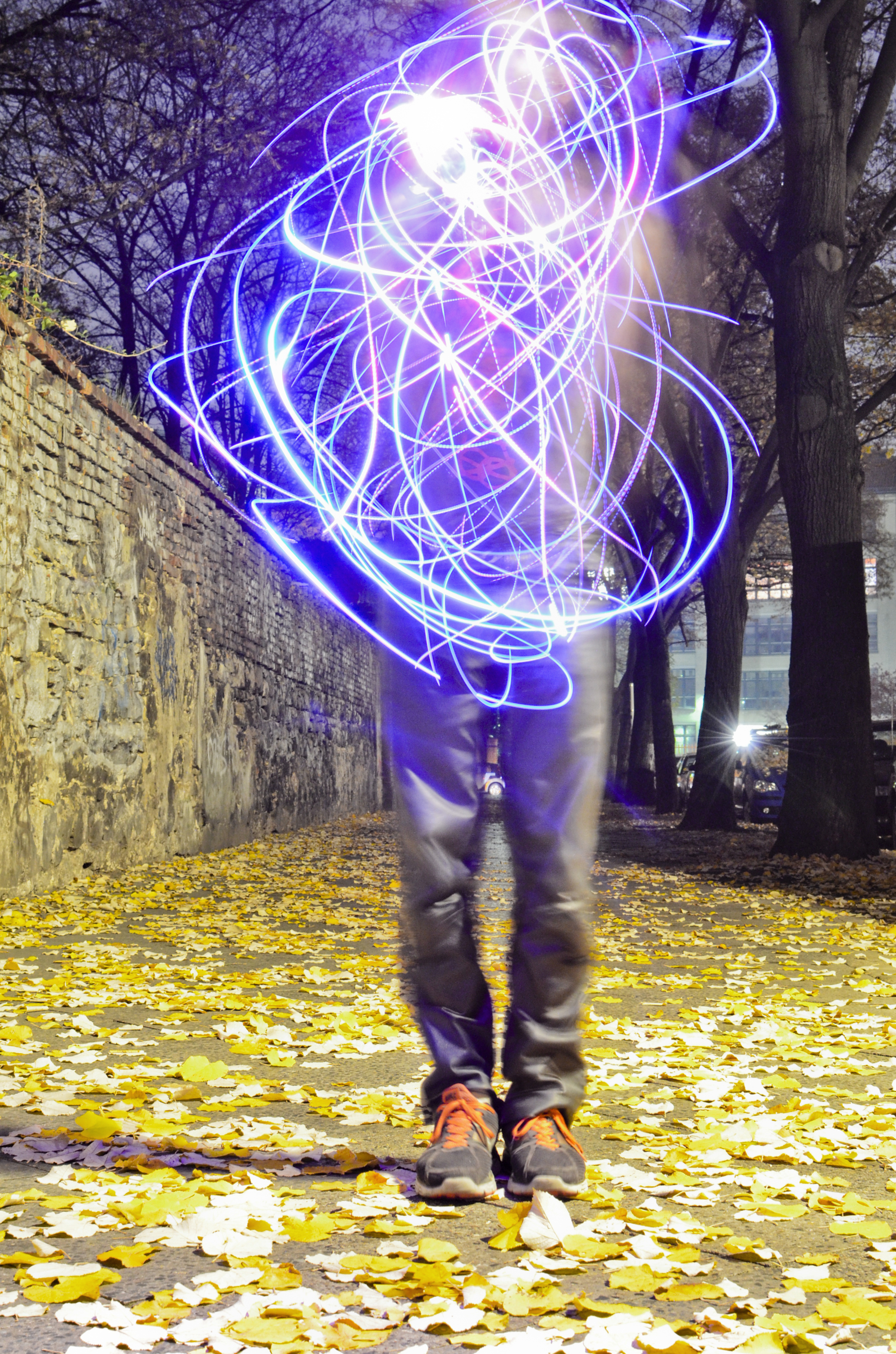 Onyx Ashanti performing outside with neon lights in the evening. Standing on a  sidewalk, on top of golden leaves that have fallen from the trees, Ashanti’s face  and torso are obscured by a tangle of blue, purple, and pink light streaks with  cloud-like light effects. Ashanti is wearing grey pants and sneakers with bright  orange laces. The street they are performing on is tree-lined and there is a  stone wall to their left.