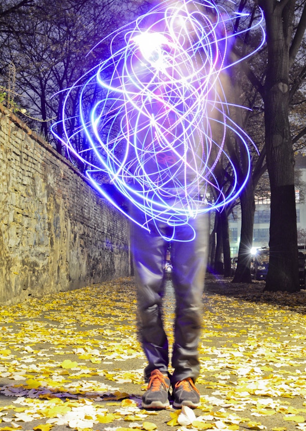 Onyx Ashanti performing outside with neon lights in the evening. Standing on a  sidewalk, on top of golden leaves that have fallen from the trees, Ashanti’s face  and torso are obscured by a tangle of blue, purple, and pink light streaks with  cloud-like light effects. Ashanti is wearing grey pants and sneakers with bright  orange laces. The street they are performing on is tree-lined and there is a  stone wall to their left.