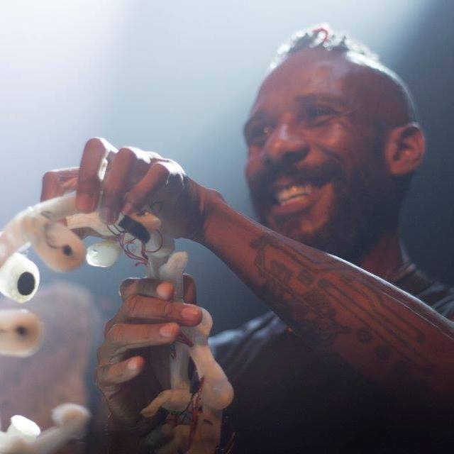 Close up of Onyx Ashanti mid-performance. Ashanti is looking out into the  audience and smiling while holding a white electronic musical interface with  red wires weaving in and out of the form. On their forearm is a circuit board-like  tattoo and they are wearing a black t-shirt. The space to Ashanti’s right is  illuminated by a bright spotlight.