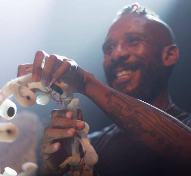 Close up of Onyx Ashanti mid-performance. Ashanti is looking out into the  audience and smiling while holding a white electronic musical interface with  red wires weaving in and out of the form. On their forearm is a circuit board-like  tattoo and they are wearing a black t-shirt. The space to Ashanti’s right is  illuminated by a bright spotlight.