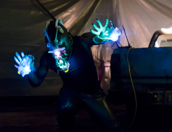 Onyx Ashanti performing in a warehouse space. Ashanti is wearing illuminated electronic musical interfaces on their face and hands, which emit blue, green, and pink lights light. Their arms are outstretched to their sides and they are looking down. Behind them hangs a yellow wire from what appears to be a mixing station. In the background are the drapes of a tent, illuminated by soft pink light.