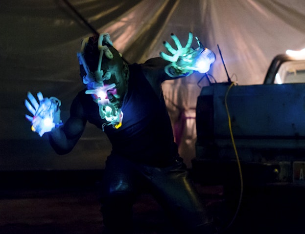 Onyx Ashanti performing in a warehouse space. Ashanti is wearing illuminated electronic musical interfaces on their face and hands, which emit blue, green, and pink lights light. Their arms are outstretched to their sides and they are looking down. Behind them hangs a yellow wire from what appears to be a mixing station. In the background are the drapes of a tent, illuminated by soft pink light.