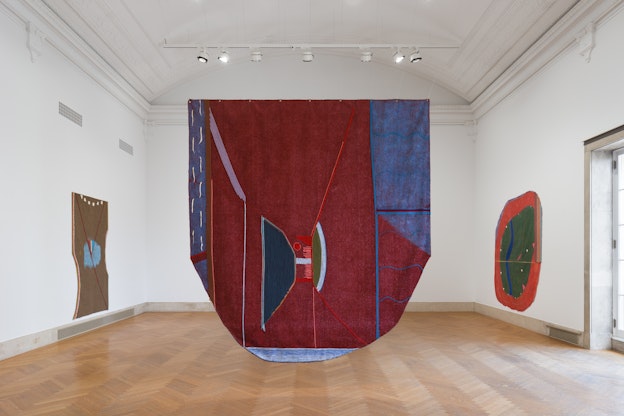 Installation view of three large paintings in a triangle formation in a gallery space. The largest of the three is shaped like a shield and acts as the top point of the triangle, suspended from the ceiling in the center of the room. It is made of blue astroturf delineated with colorful yarn and paint, which form a variety of shapes, and seed pods that are stitched to the surface. It is mainly a deep red, which contrasts against the white walls of the room, but also includes violet, dark blue, orange, green, baby blue, and white. The other paintings are installed on parallel walls. On the right wall is a painting of a round green shape with a round, thick orange border surrounding it. Inside of the green is a blue squiggly shape that resembles a lake, as well as a number of pink lines and white and pink triangles spread throughout. There are also small lines and shapes on the orange border, including a blue squiggly line and four rectangular shapes stiched together, in red, white, yellow, and blue. On the opposite wall is an organic rectangular shape, predominantly in brown with a blue round shape in the center of it. A red “V” begins in each of the top corners and extends to the very middle of the blue, at which point the “V” turns into a red diagonal line that extends from the center of the painting to its bottom right corner.